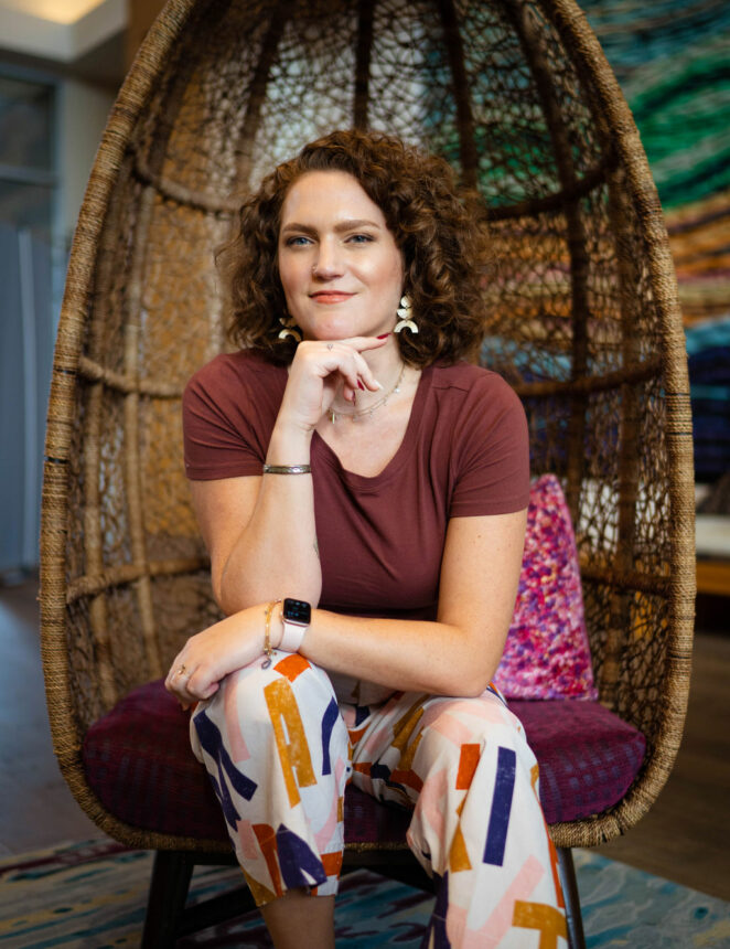 CEO of Lyon Content writing agency, Christina Lyon, sits in an egg shaped wicker chair with her hands rested on her knee and beneath her chin, wearing colorful pants and a brown t-shirt.