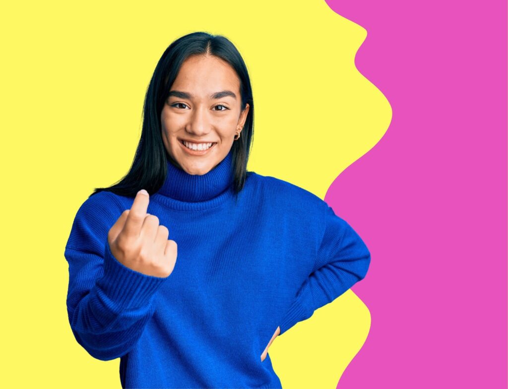 Young woman in blue turtleneck sweater smiling and making "come here" hand motion in front of yellow and pink background