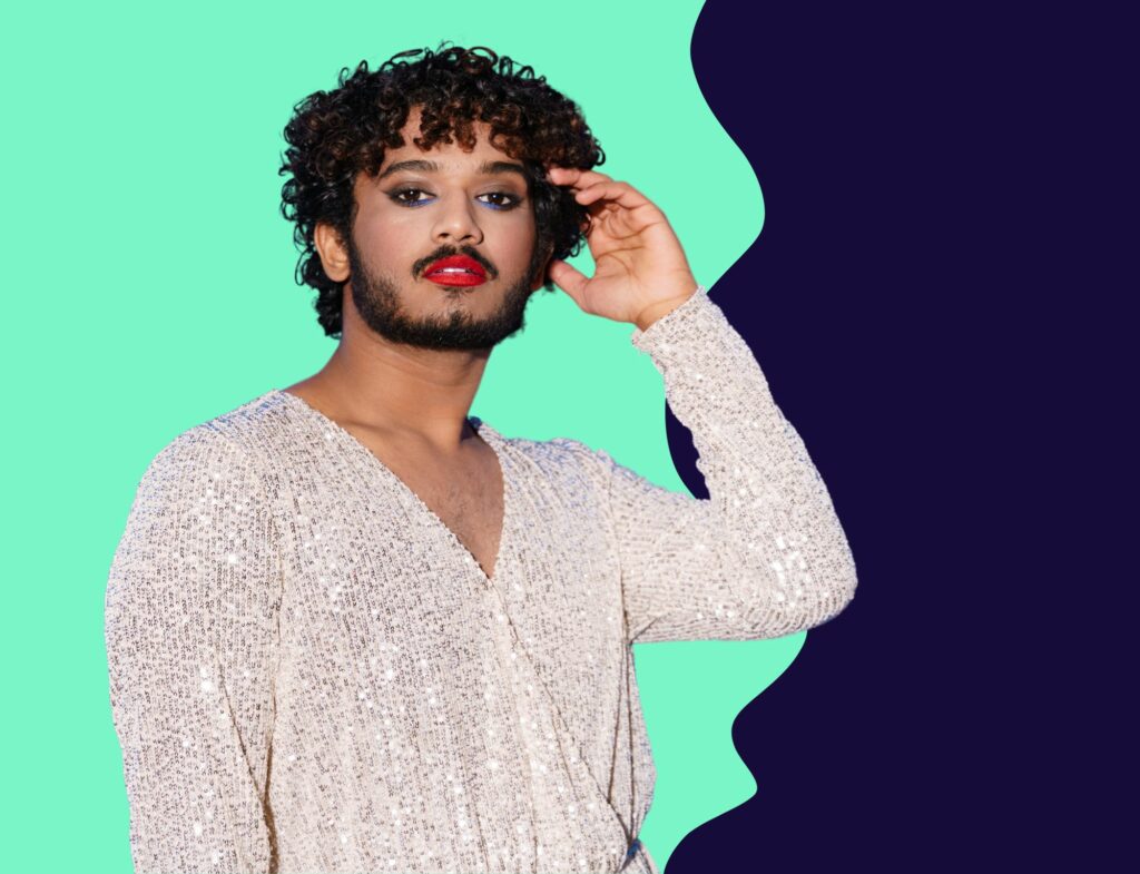 Male presenting model wearing makeup and sparkling shirt in front of green and navy background